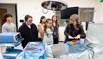 A female healthcare professional is explaining a surgical training mannequin to a group of five students. The mannequin is covered in a gray sheet, revealing a simulated human torso with an orange representing an organ. Surgical instruments and equipment, including a ventilator, are visible on a nearby table. The setting appears to be a medical training room.